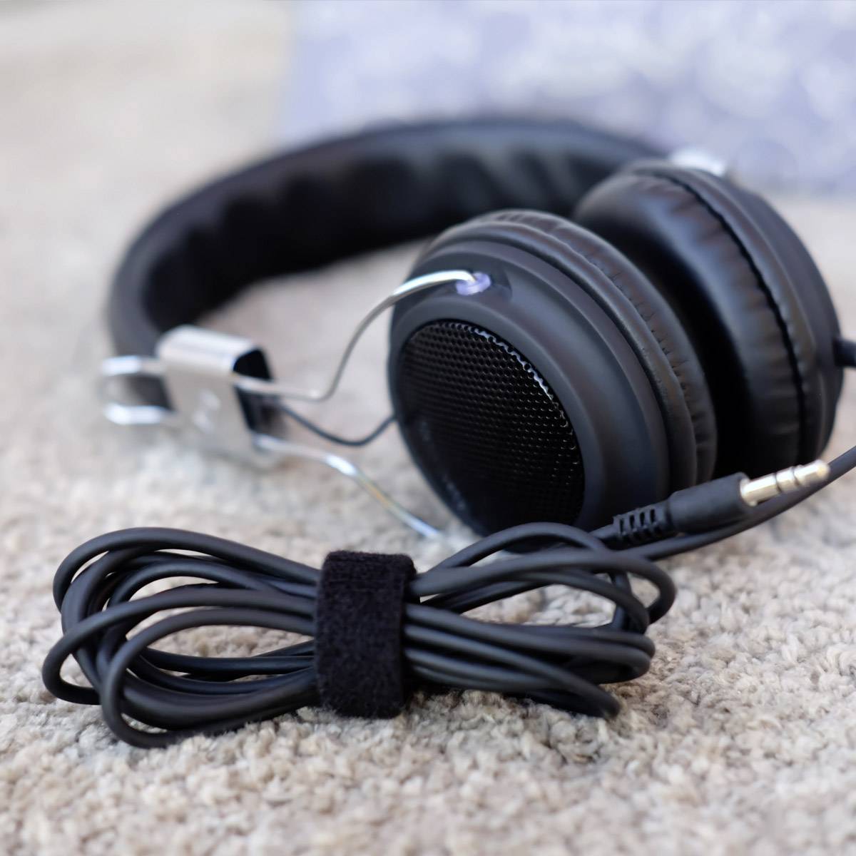 Schwarze Over-Ear-Kopfhörer mit gepolsterten Ohrmuscheln liegen auf einem beigen Teppich, das Kabel ist ordentlich aufgerollt.