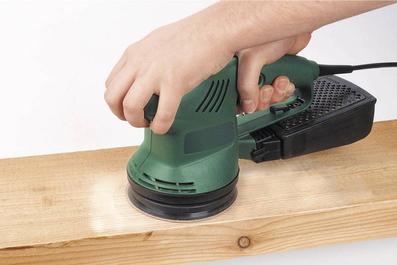 Eine Hand benutzt einen grünen Schwingschleifer, um ein Stück Holz zu bearbeiten. Der Schleifer wird über die Holzoberfläche geführt.