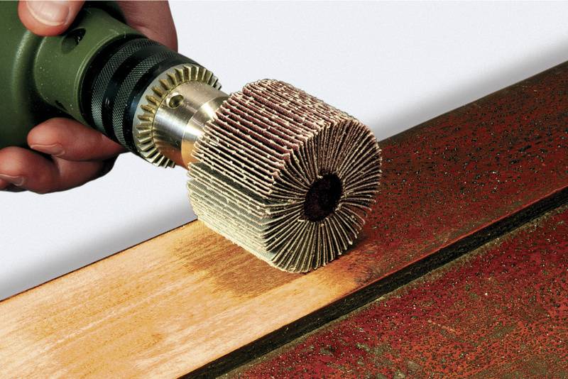 Eine Hand hält ein elektrisches Schleifgerät, das eine Holzfläche bearbeitet. Die Schleifscheibe ist zylindrisch und wird für präzise Arbeiten verwendet.