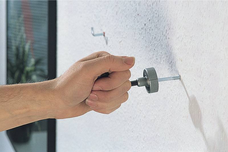 Eine Hand dreht mit einem Werkzeug eine große Schraube in eine weiße Wand. Im Hintergrund ist eine Pflanze zu sehen.