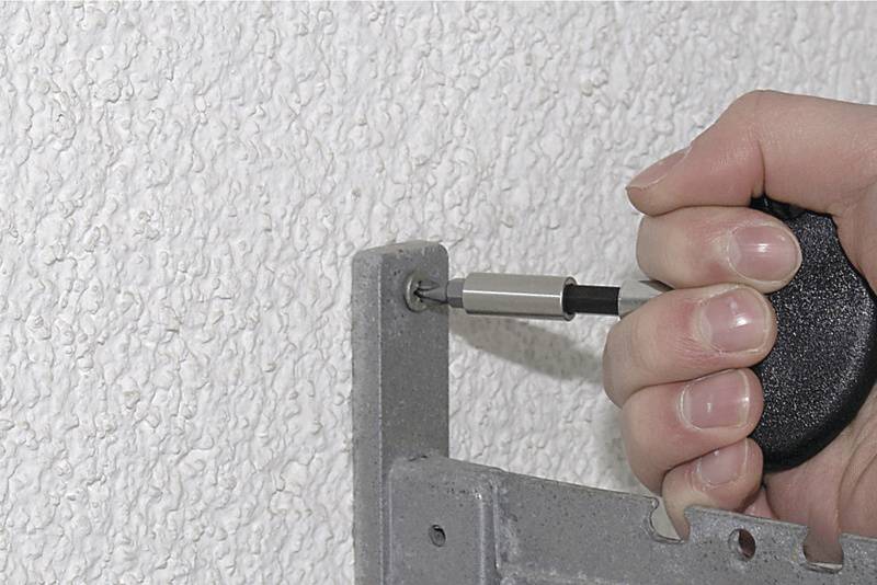 Eine Hand zieht mit einem Schraubendreher eine Schraube in eine Metallschiene, die an einer weißen, strukturierten Wand befestigt wird.