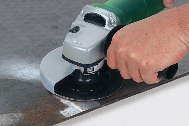 Eine Hand hält einen Winkelschleifer, der auf einer Metalloberfläche arbeitet. Funken oder Staubpartikel sind nicht sichtbar.