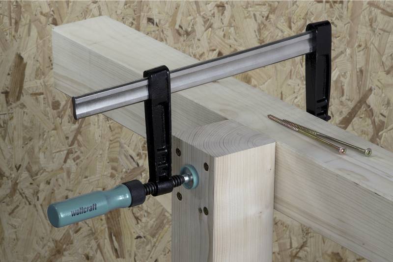 Eine Zwinge klemmt zwei Holzbalken zusammen. Ein Schraubendreher und eine Schraube liegen daneben auf einem Holzuntergrund.