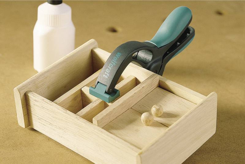 Eine Holzkiste mit Trennwänden wird mit einer Zwinge geleimt. Daneben steht eine Flasche Leim.