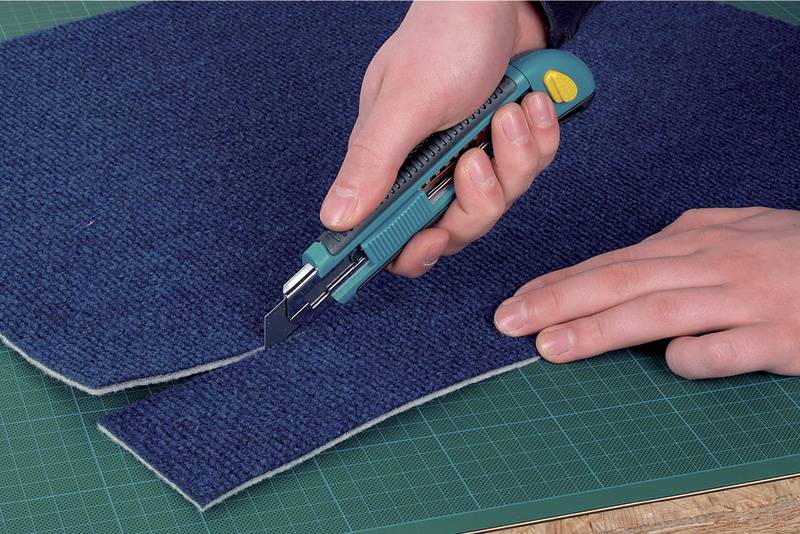 Eine Person schneidet einen blauen Teppich mit einem grünen Cuttermesser auf einer Schneidematte.