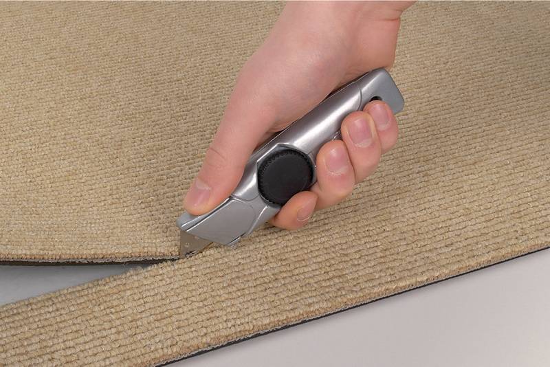 Hand schneidet mit Teppichmesser einen beigen Teppich auf weißer Oberfläche.