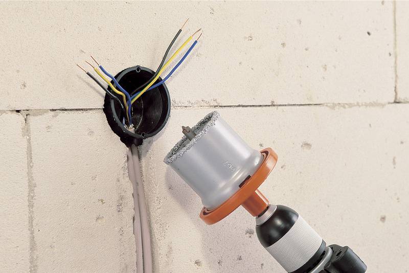 Gipswand mit offenem Installationsdose und abisolierten Kabeln; daneben ein Bohrer mit Lochsägeaufsatz, der zum Bohren verwendet wird.