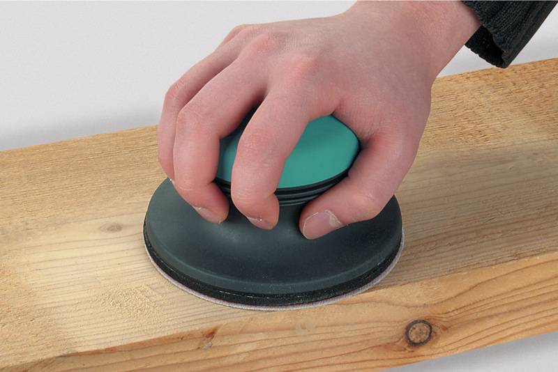 Eine Hand schleift mit einem runden Schleifblock ein Holzbrett. Der Schleifblock hat eine grüne Oberseite.