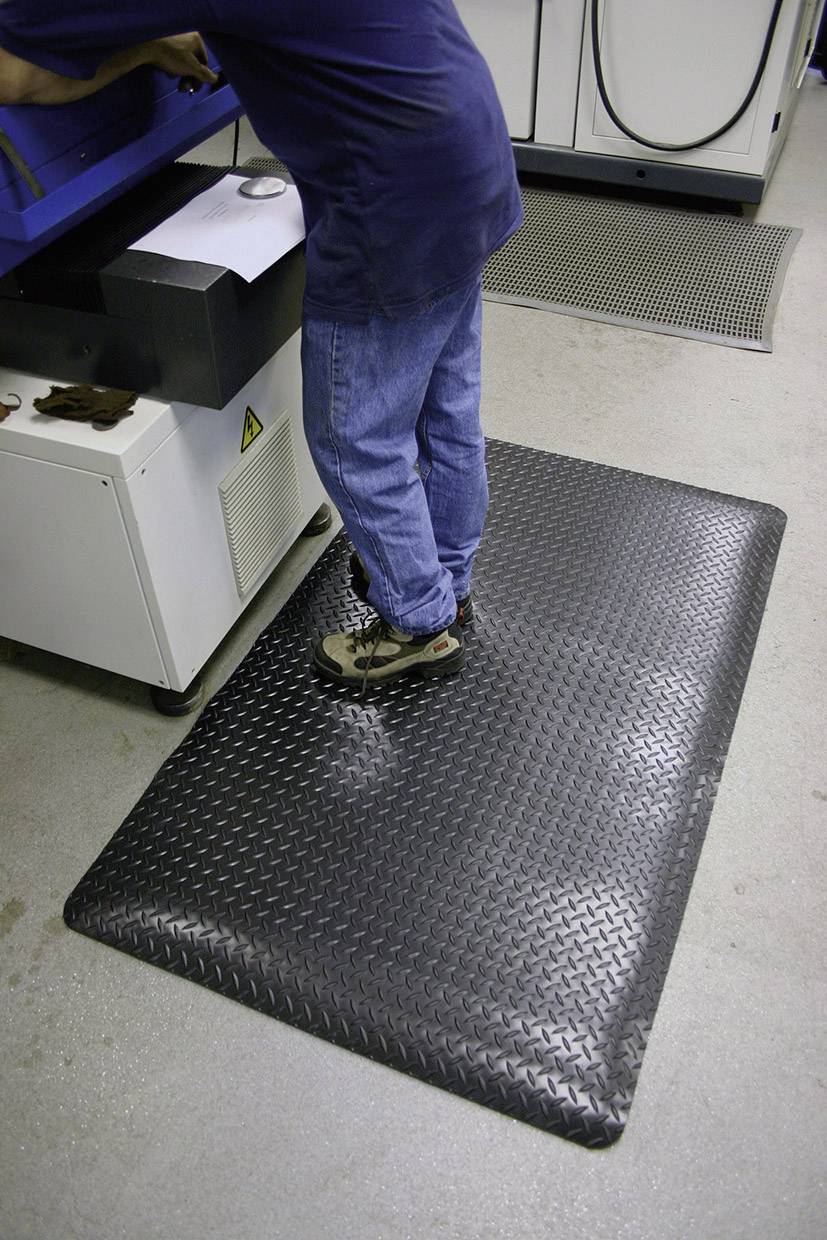 Person steht auf einer gepolsterten schwarzen Matte vor einer Maschine in einer Werkstattumgebung.