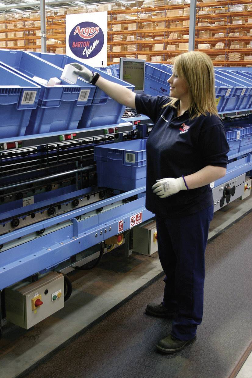 Eine Frau in Arbeitskleidung sortiert Pakete in blauen Kisten auf einem Förderband in einem Lager. Im Hintergrund sind Regale mit Kartons zu sehen.