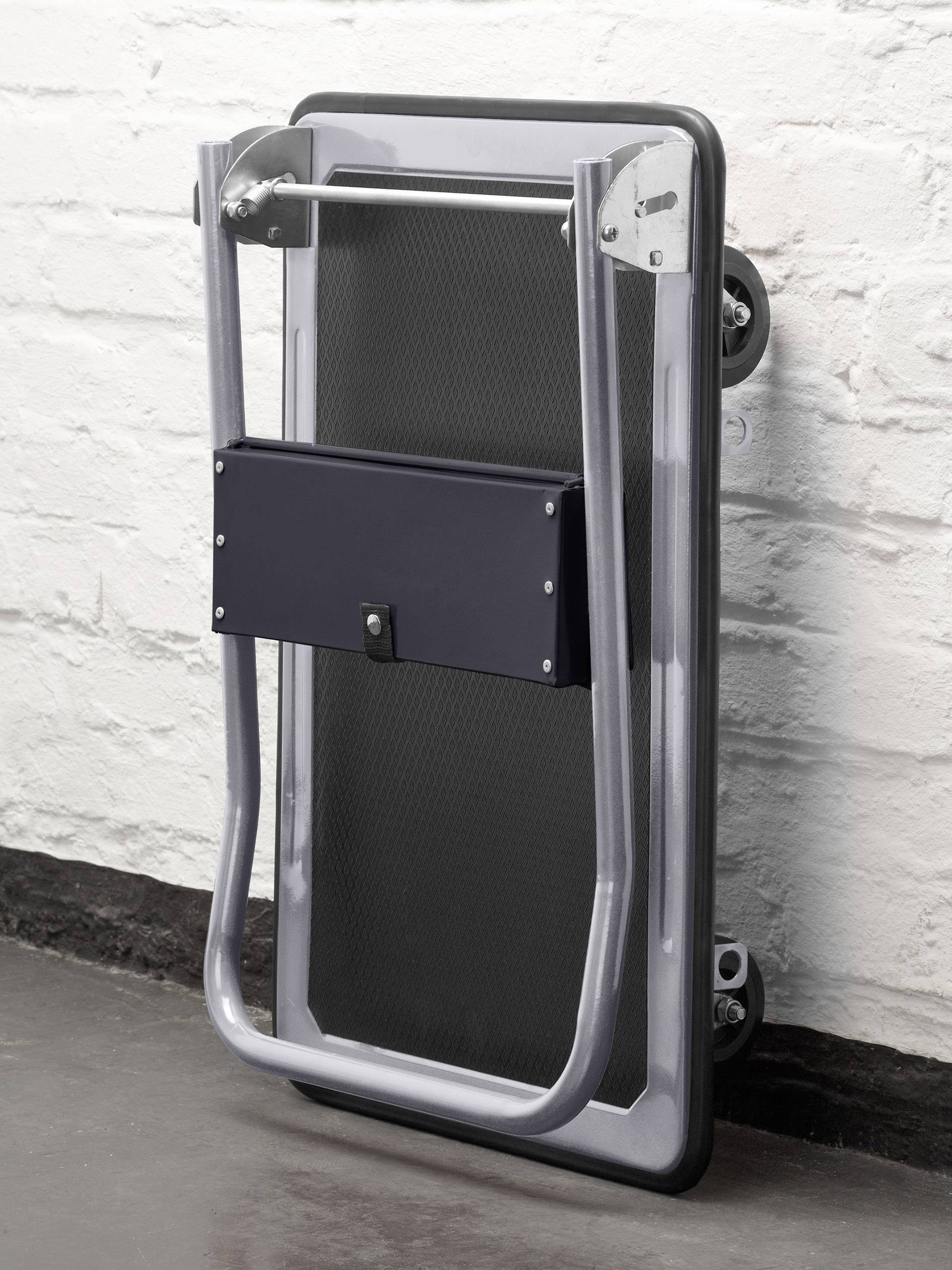 Zusammengeklappter schwarzer Rollator steht an einer weißen Wand. Er hat ein silbernes Gestell und schwarze Räder.