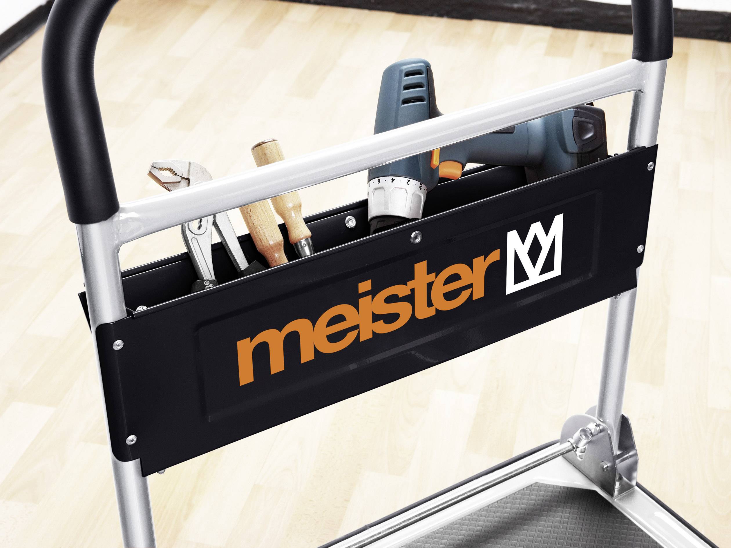 Werkzeugtasche auf einem Trolley, mit Schraubenzieher, Zange, Hammer und Bohrmaschine, vor hellem Holzfußboden. Aufschrift: 'meister'.