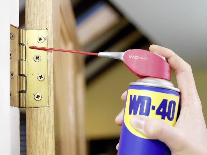 Eine Hand sprüht Öl aus einer WD-40-Dose auf ein Türscharnier, um es zu schmieren.