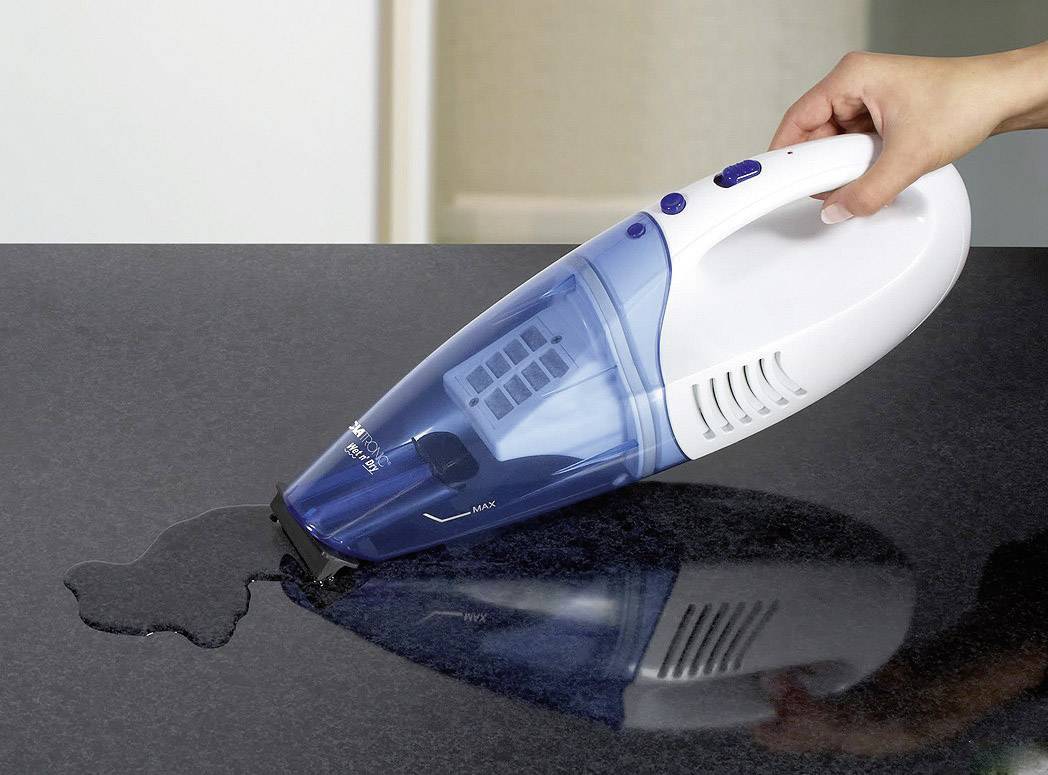 Eine Hand hält einen blauen Handstaubsauger, der verschüttete Flüssigkeit von einer glatten, dunklen Oberfläche aufsaugt.