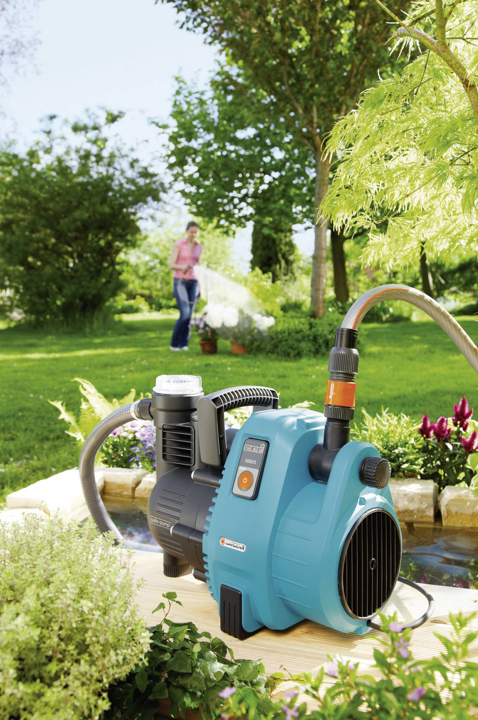 Eine blaue Gartenpumpe mit digitalem Display und Schläuchen auf einem Tisch im Garten. Im Hintergrund gießt eine Person Pflanzen.