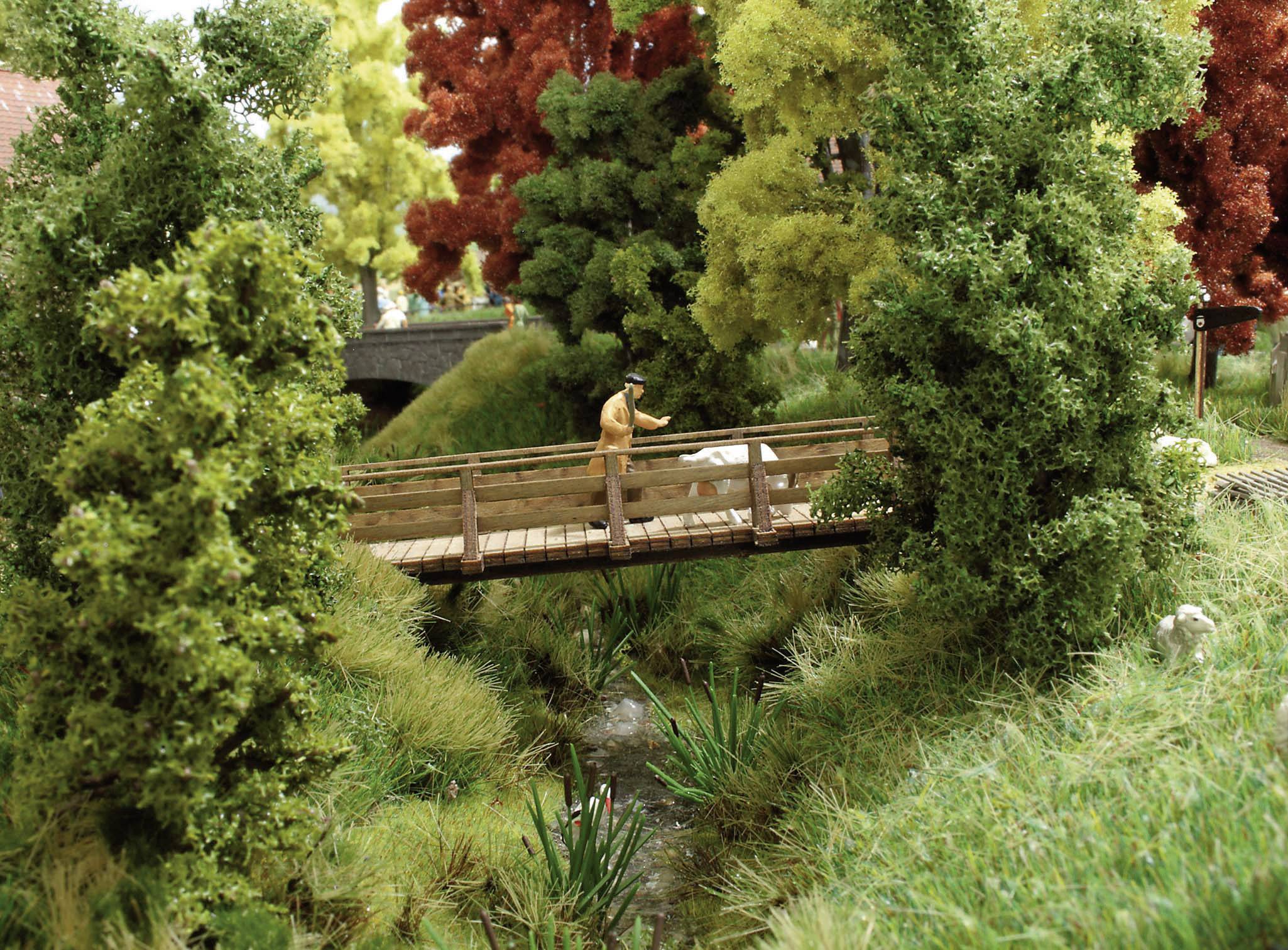 Ein Modell eines Mannes auf einer Holzbrücke in einem Miniaturpark, umgeben von bunten Bäumen und einem kleinen Bach darunter.
