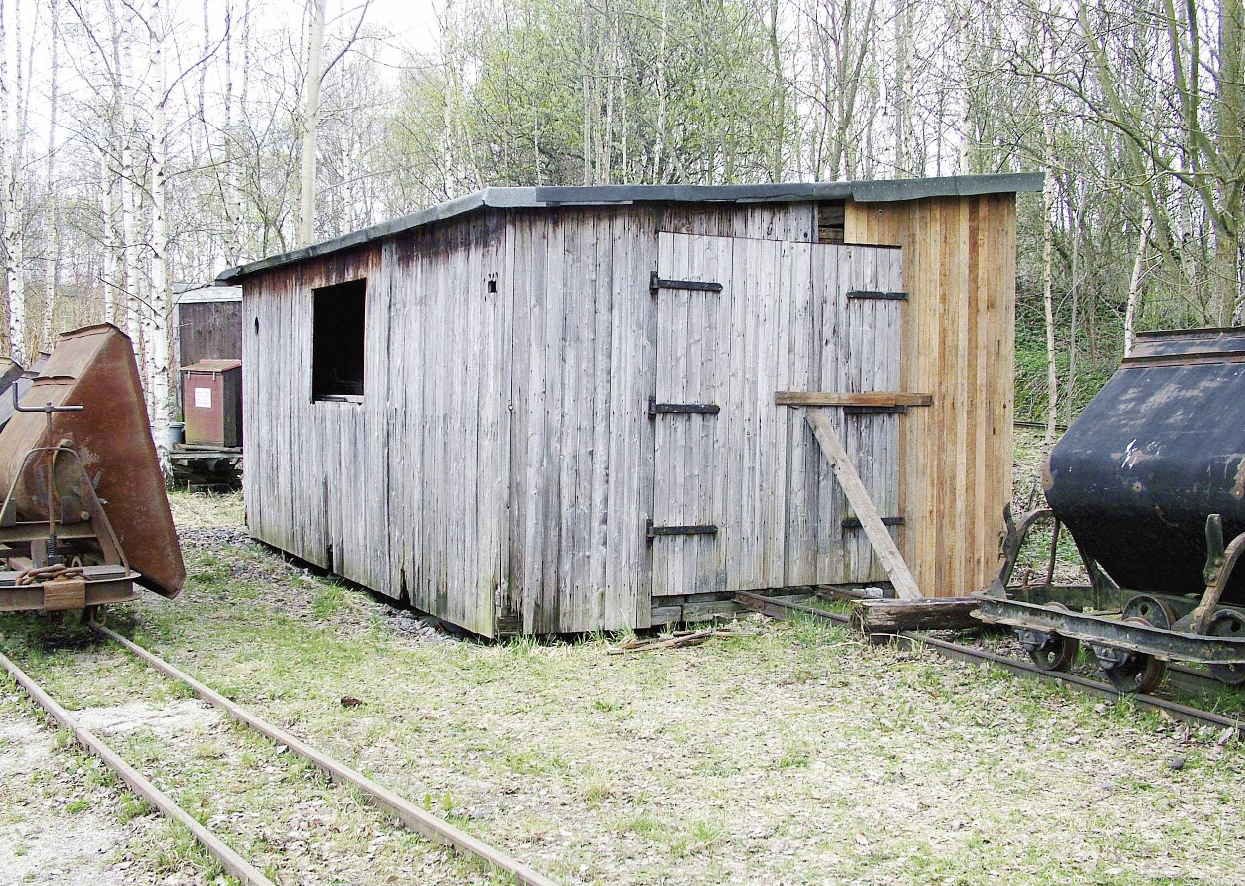 Holzhütte neben einer alten Eisenbahnstrecke, umgeben von Bäumen. Links ein rostiger Waggon, rechts alte Kohlenanhänger.