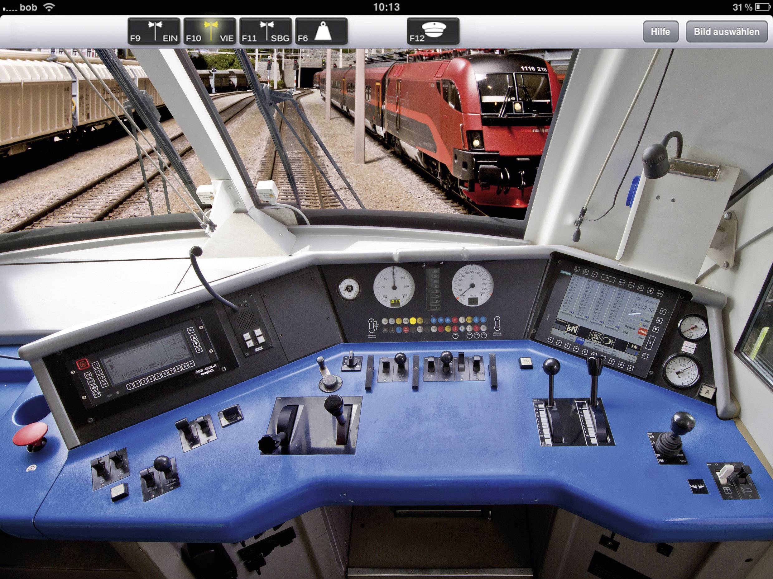 Fahrerstand eines Zuges: verschiedene Bedienelemente und Anzeigen, im Hintergrund rote Lokomotive auf den Gleisen sichtbar.