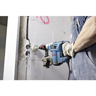Ein Arbeiter bohrt mit einem Bohrhammer Löcher in eine Wand, während freiliegende Kabel sichtbar sind.