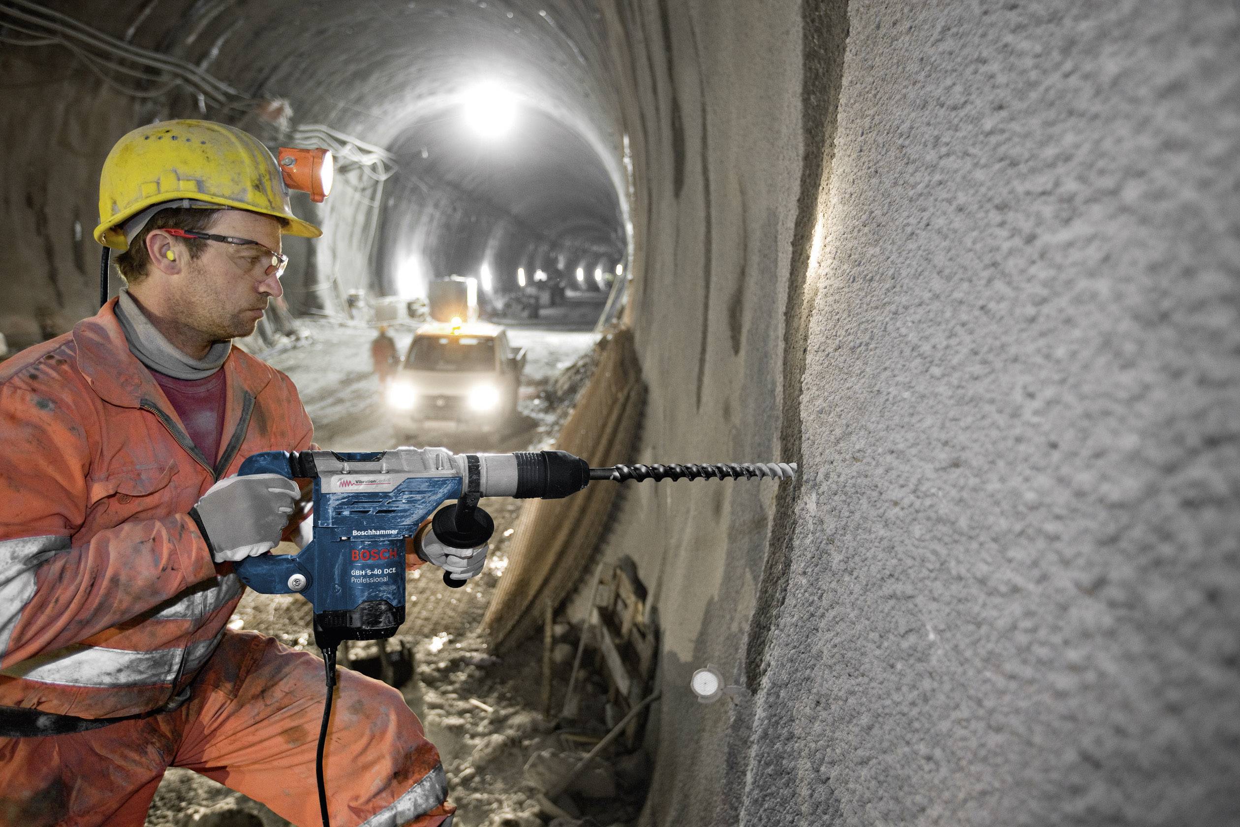 Ein Bauarbeiter in Sicherheitskleidung verwendet eine Bohrmaschine in einem Tunnel, während im Hintergrund ein Fahrzeug sichtbar ist.