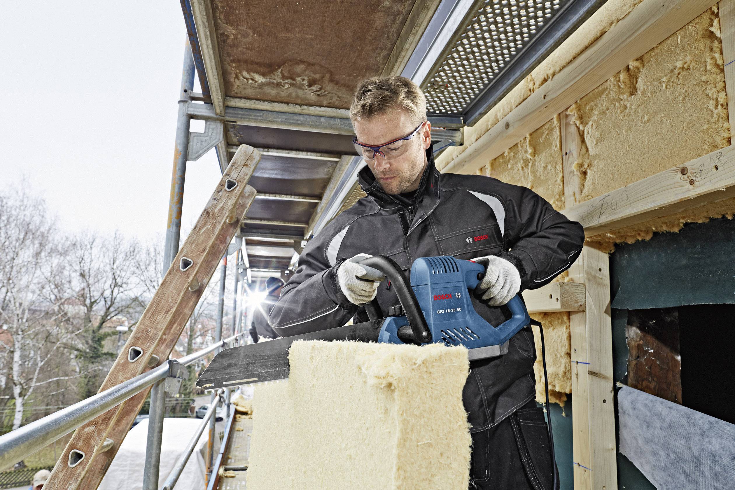 Ein Arbeiter auf einem Gerüst schneidet mit einem Elektrowerkzeug Dämmmaterial an einem Gebäude. Schutzbrille und Handschuhe sind getragen.