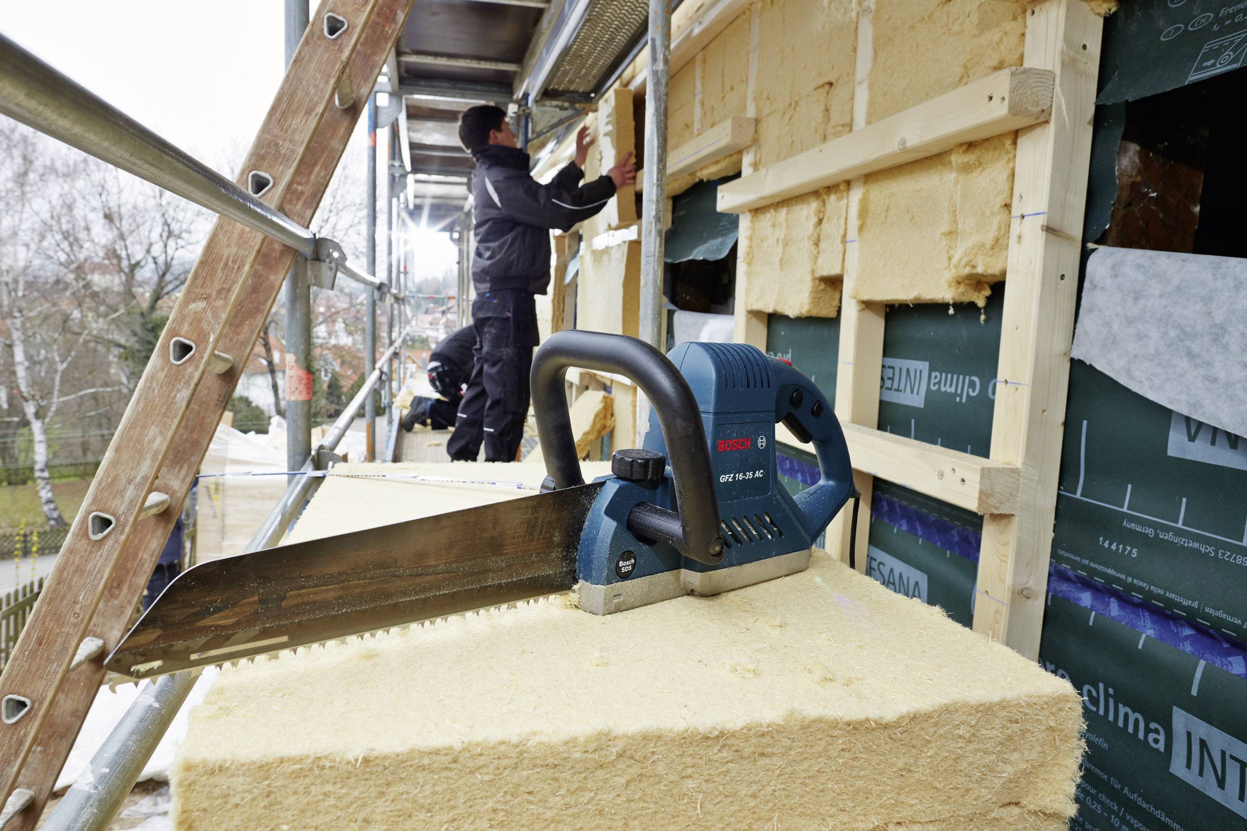 'Ein Arbeiter isoliert die Fassade eines Gebäudes mit Dämmmaterial, während im Vordergrund eine elektrische Säge liegt.'