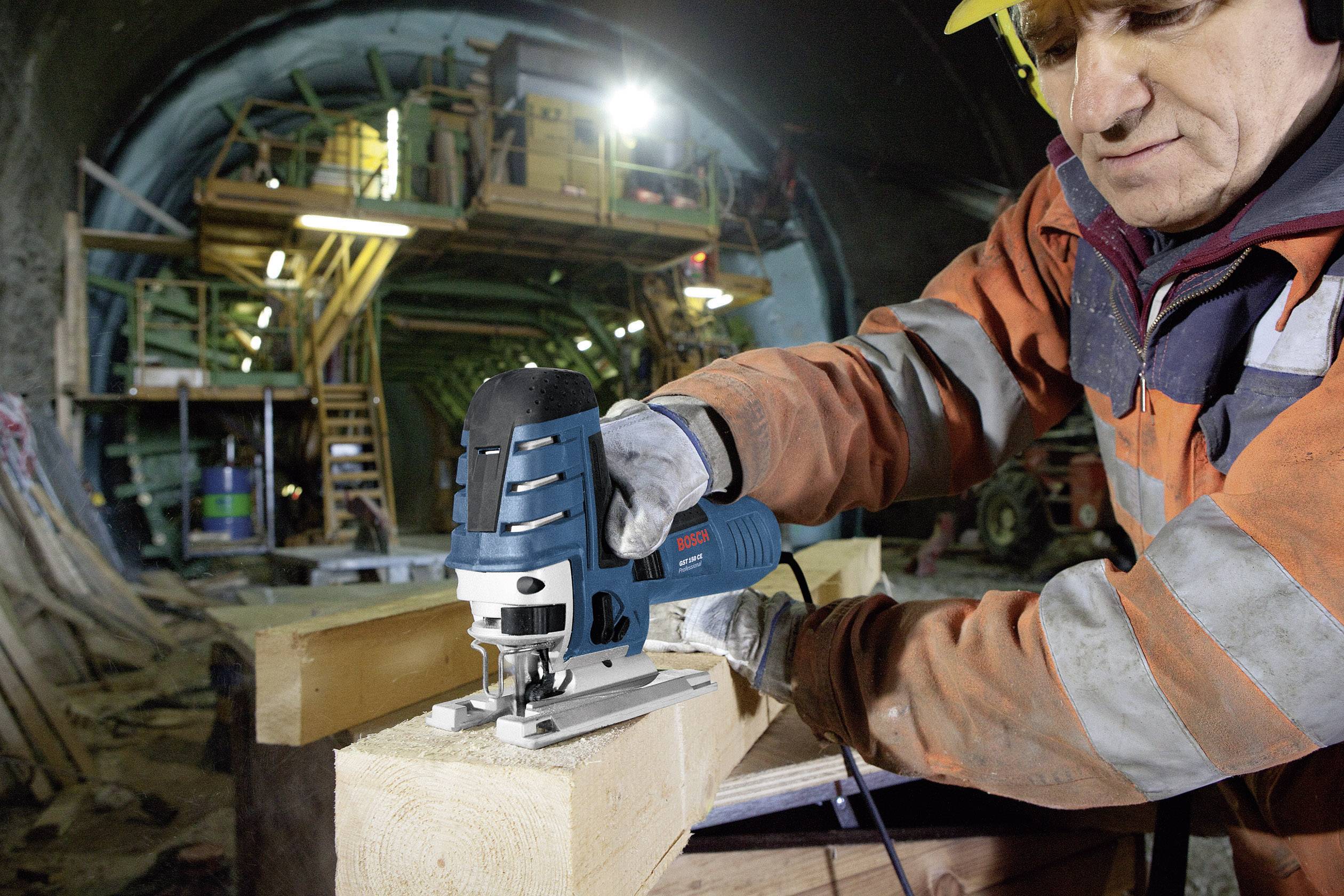 Ein Bauarbeiter trägt Schutzkleidung und nutzt eine elektrische Stichsäge, um ein Brett zu schneiden. Im Hintergrund ist eine Baustelle im Tunnel zu sehen.