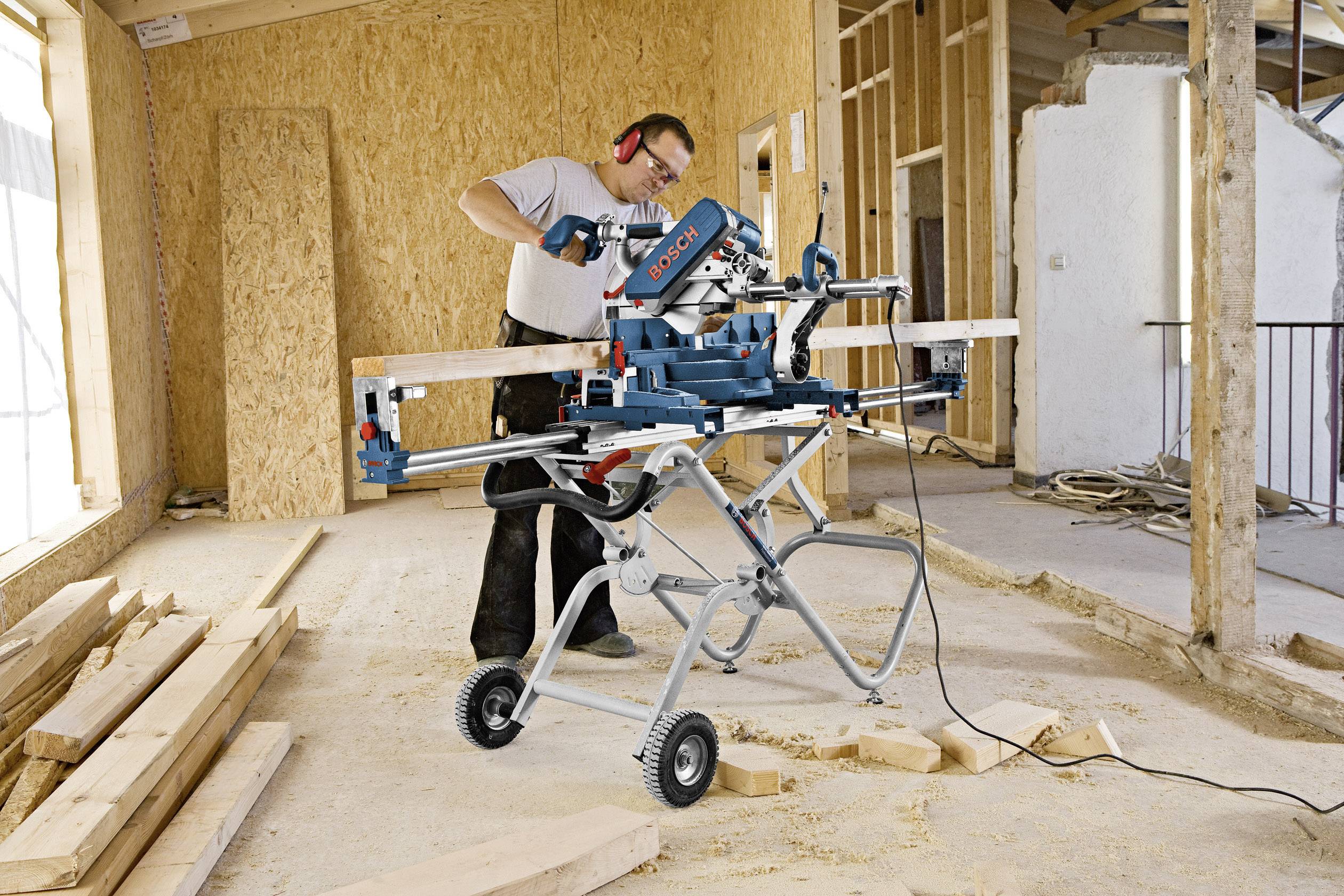 Ein Bauarbeiter verwendet eine elektrische Gehrungssäge auf einer Baustelle, um Holz zu schneiden. Im Hintergrund sind Wände in Bauweise erkennbar.