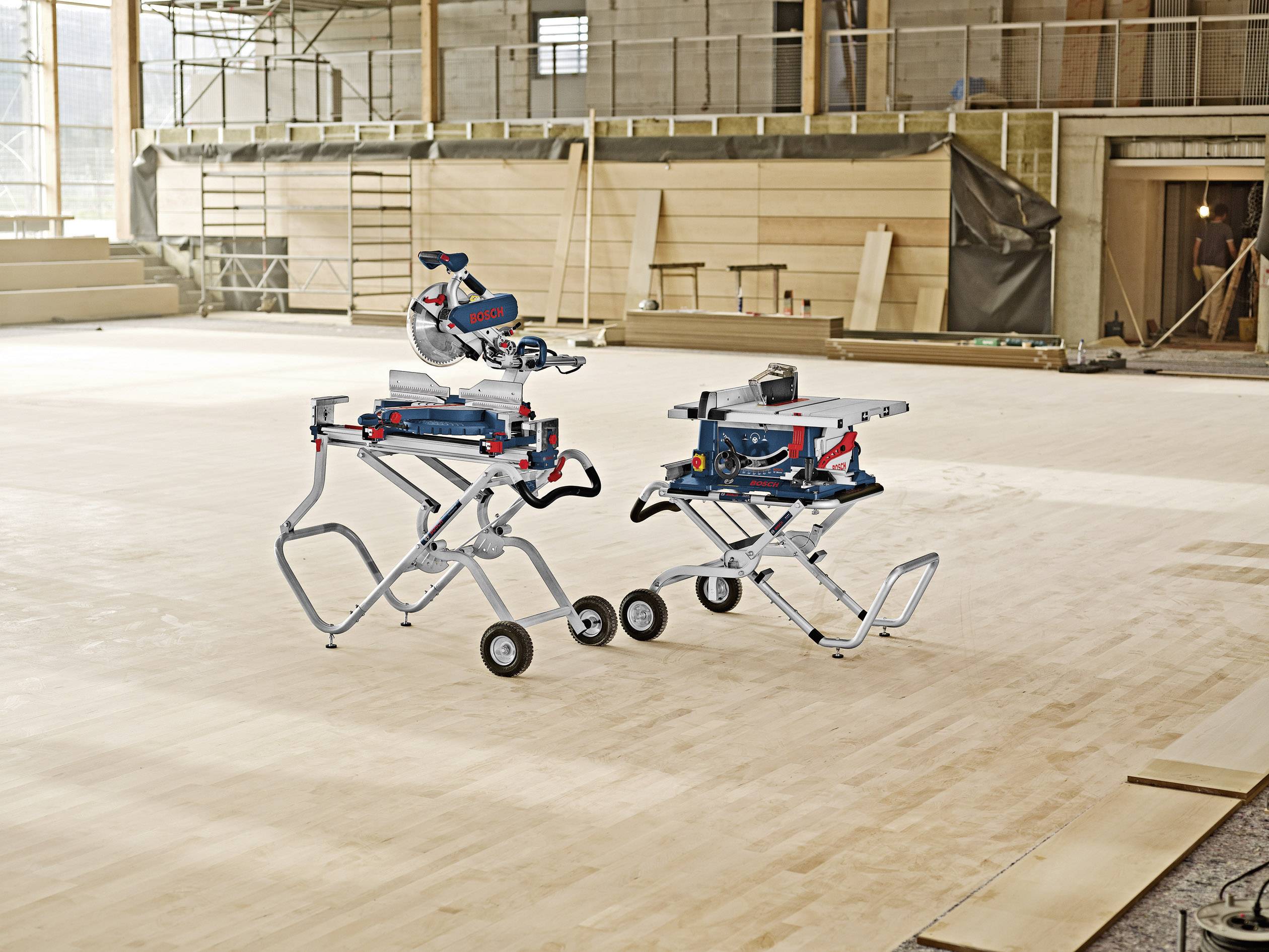 Zwei elektrische Werkzeuge auf mobilen Ständern, eine Tischsäge und eine Gehrungssäge, stehen auf einem Holzboden in einer Baustellenumgebung.