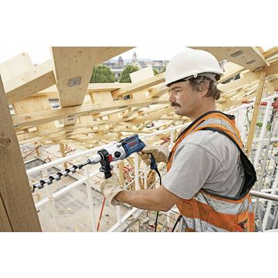 Ein Bauarbeiter in Schutzhelm und Weste arbeitet auf einer Holzbaustelle mit einem Bohrer. Im Hintergrund sind Gebäude zu sehen.