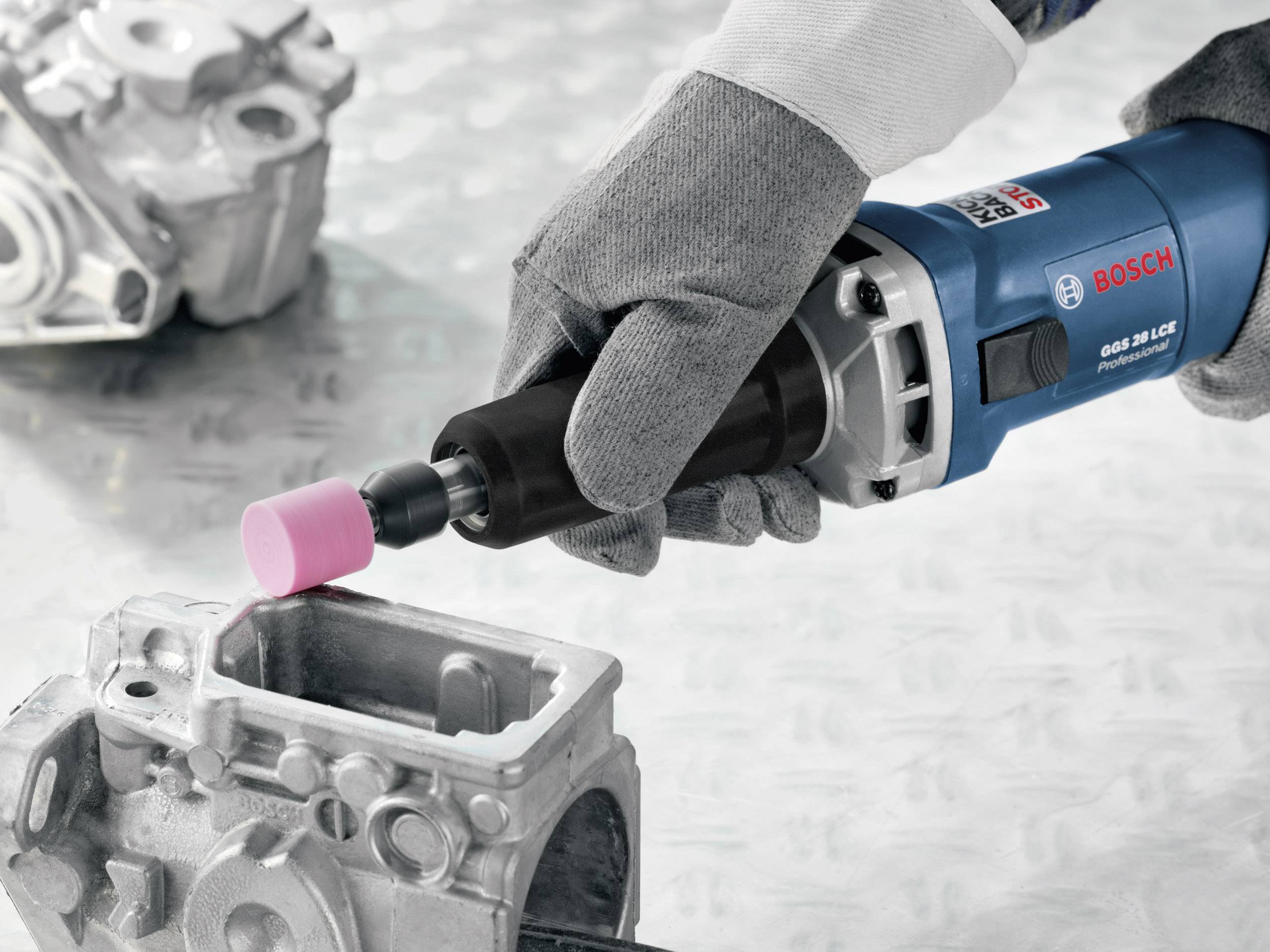 Eine Hand mit einem grauen Handschuh hält ein blaues Bosch-Werkzeug und bearbeitet ein Metallteil mit einem rosa Schleifkopf.