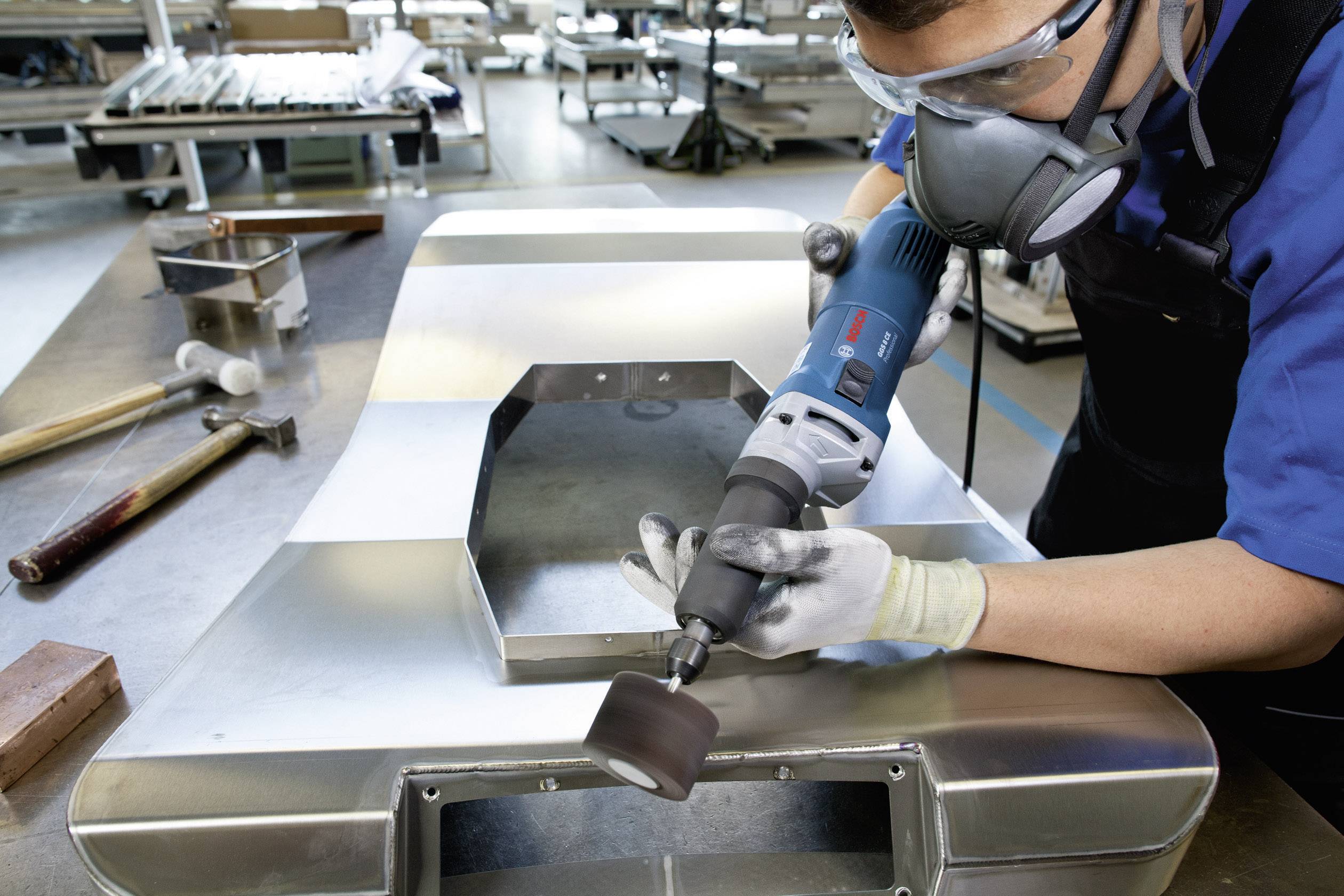 Person schleift Metalloberfläche mit einer elektrischen Schleifmaschine in einer Werkstatt, trägt Schutzbrille und Atemmaske.