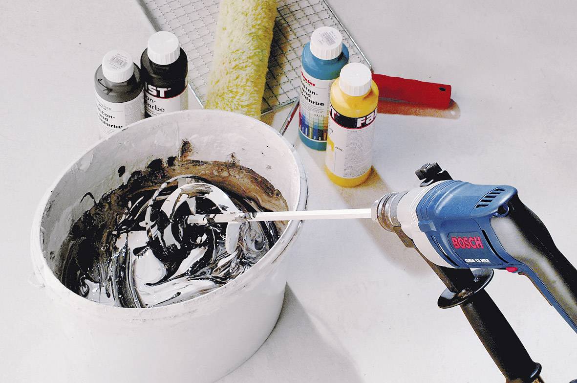 Eine Bohrmaschine von Bosch mischt Farbe in einem Eimer. Daneben stehen Farbdosen und ein Pinsel auf einem Tisch.