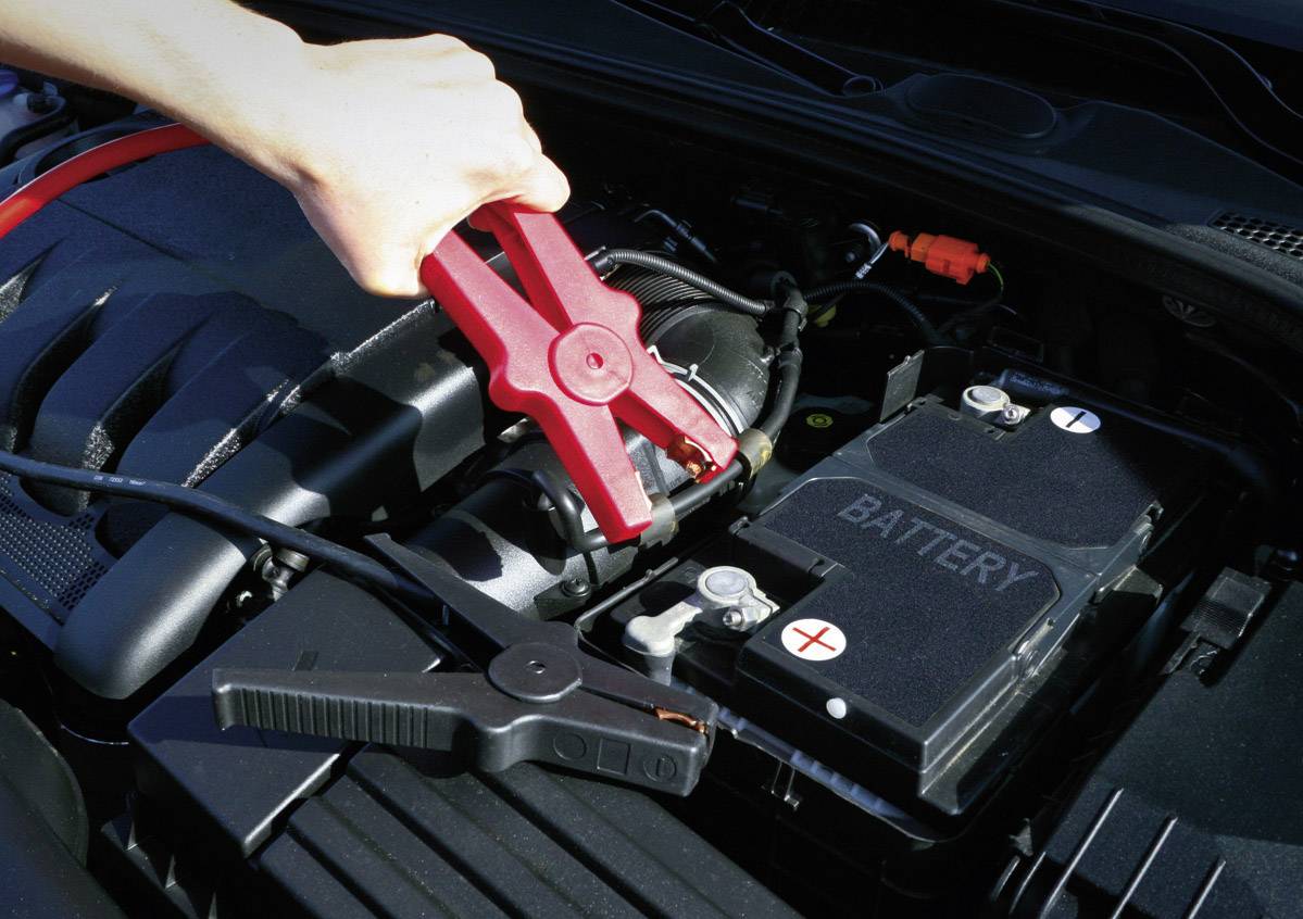 Eine Hand hält rote Starthilfekabel an einer Autobatterie, um das Fahrzeug zu starten. Die Batterie ist bereits mit den Kabeln verbunden.