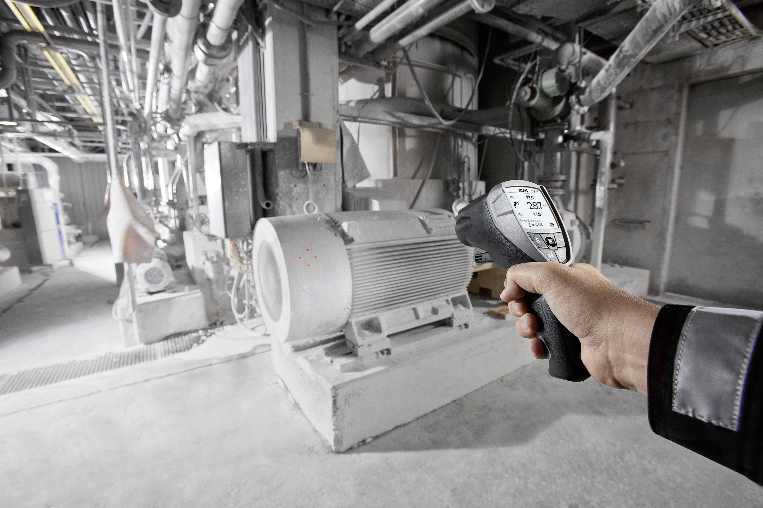 Eine Person misst die Temperatur eines großen Industriemotors mit einem Infrarot-Thermometer. Der Raum ist maschinell voll ausgestattet.