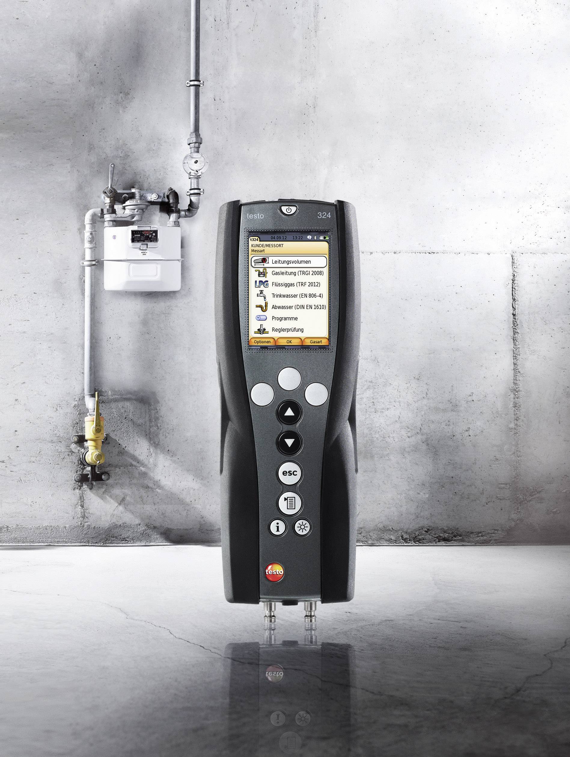 Ein tragbares Abgasmessgerät steht auf einem Boden vor einer grauen Wand in einem industriellen Raum. Im Hintergrund ist ein Gaszähler.