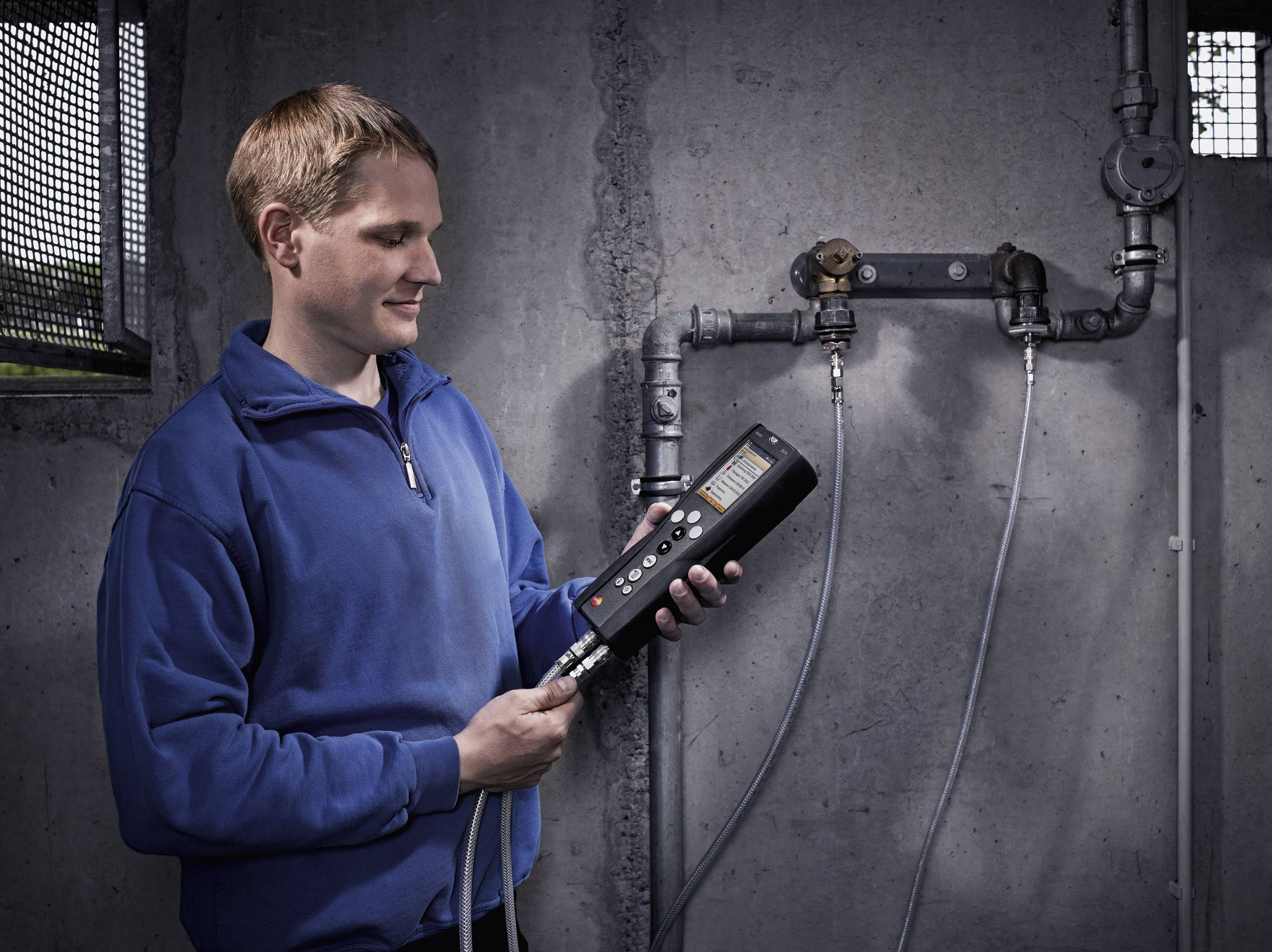 Eine Person in blauer Jacke bedient ein tragbares Messgerät, angeschlossen an Rohre an einer Betonwand, möglicherweise zur Lecksuche.