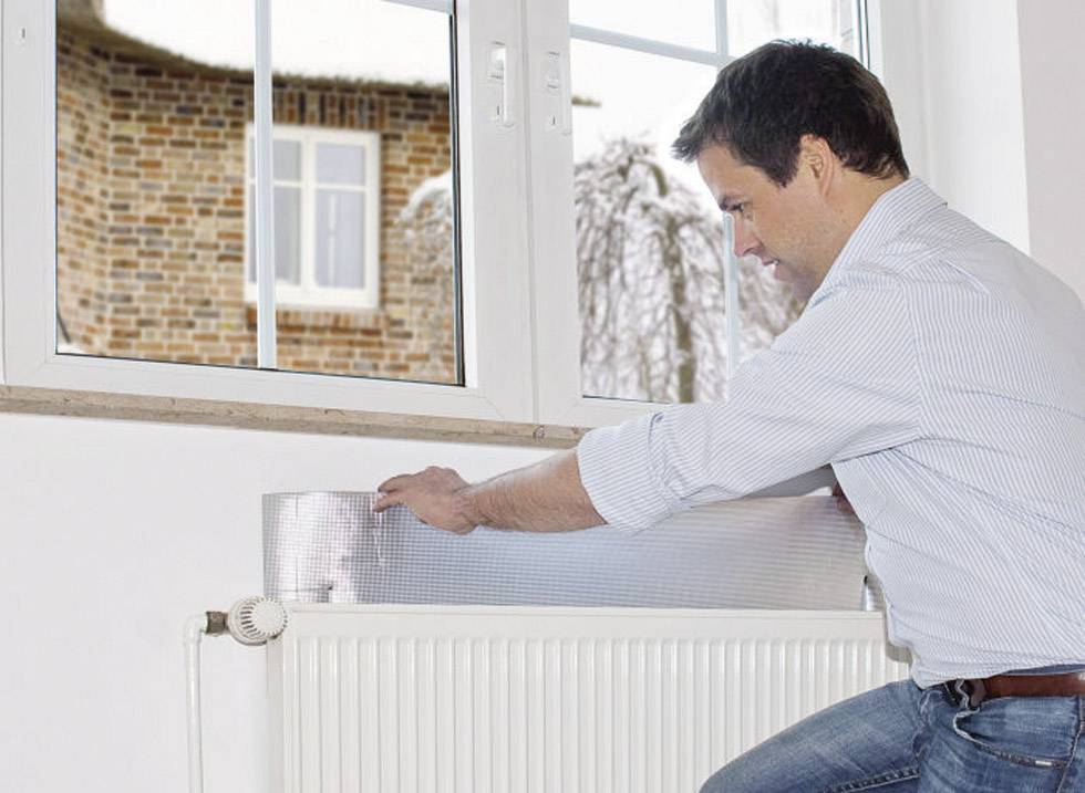Ein Mann befestigt eine Isolierfolie hinter einem Heizkörper, um Energieeffizienz zu verbessern. Ein Fenster zeigt eine verschneite Landschaft.