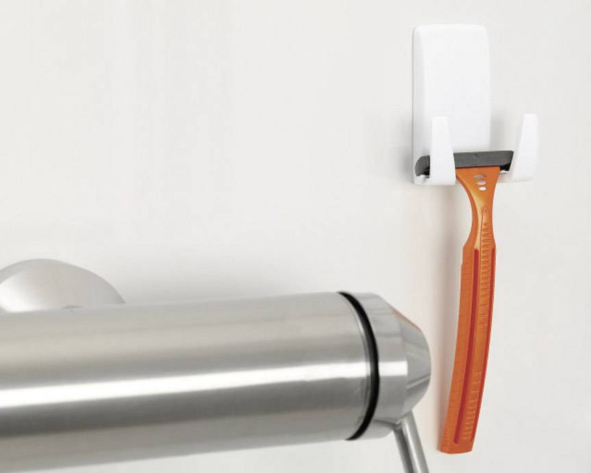 Ein orangefarbener Rasierer hängt an einem weißen Wandhalter neben einem verchromten Handtuchhalter.