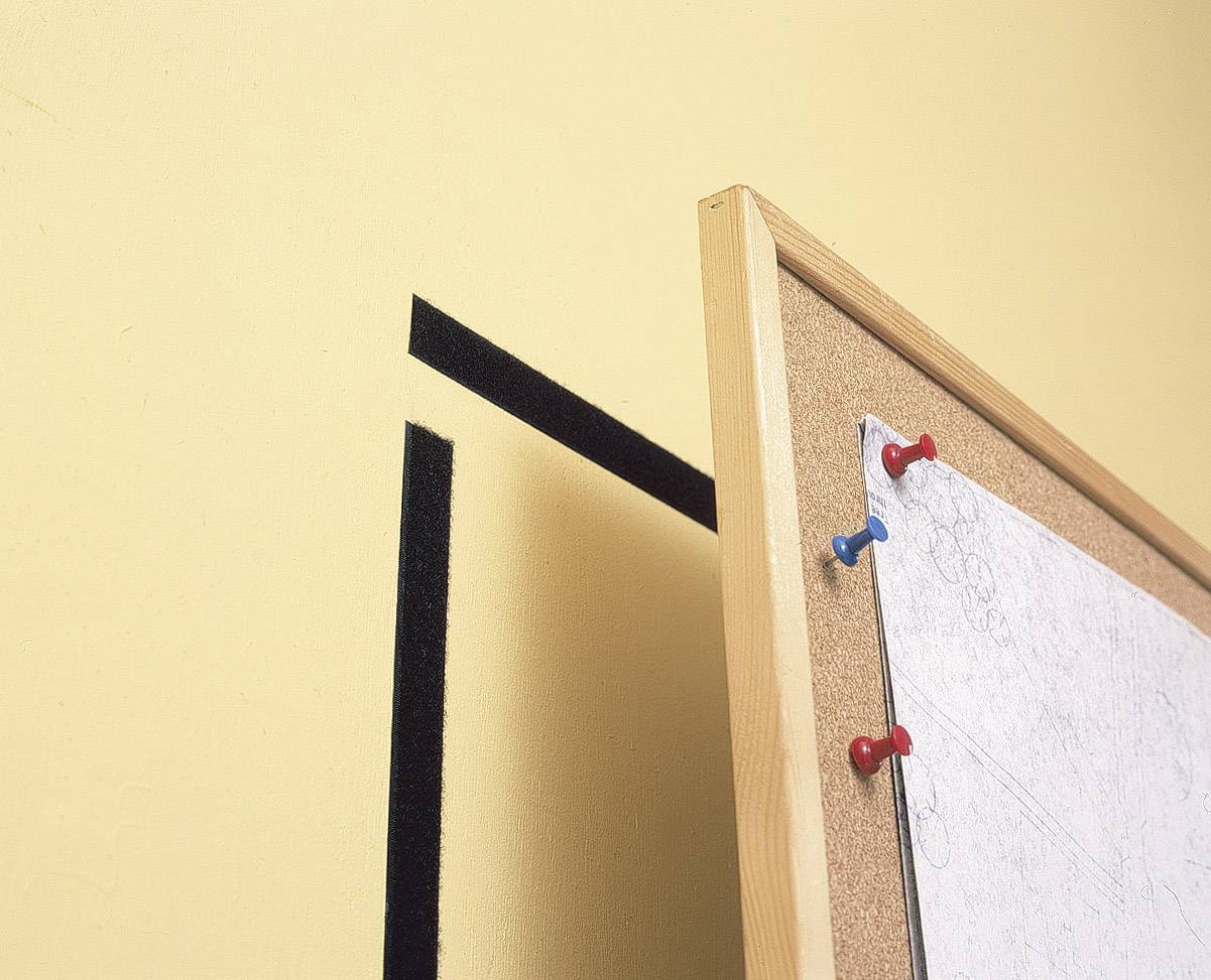 Ein Korkbrett mit weißen Papieren, befestigt mit bunten Stecknadeln, lehnt an einer gelben Wand mit schwarzem Liniendetail.