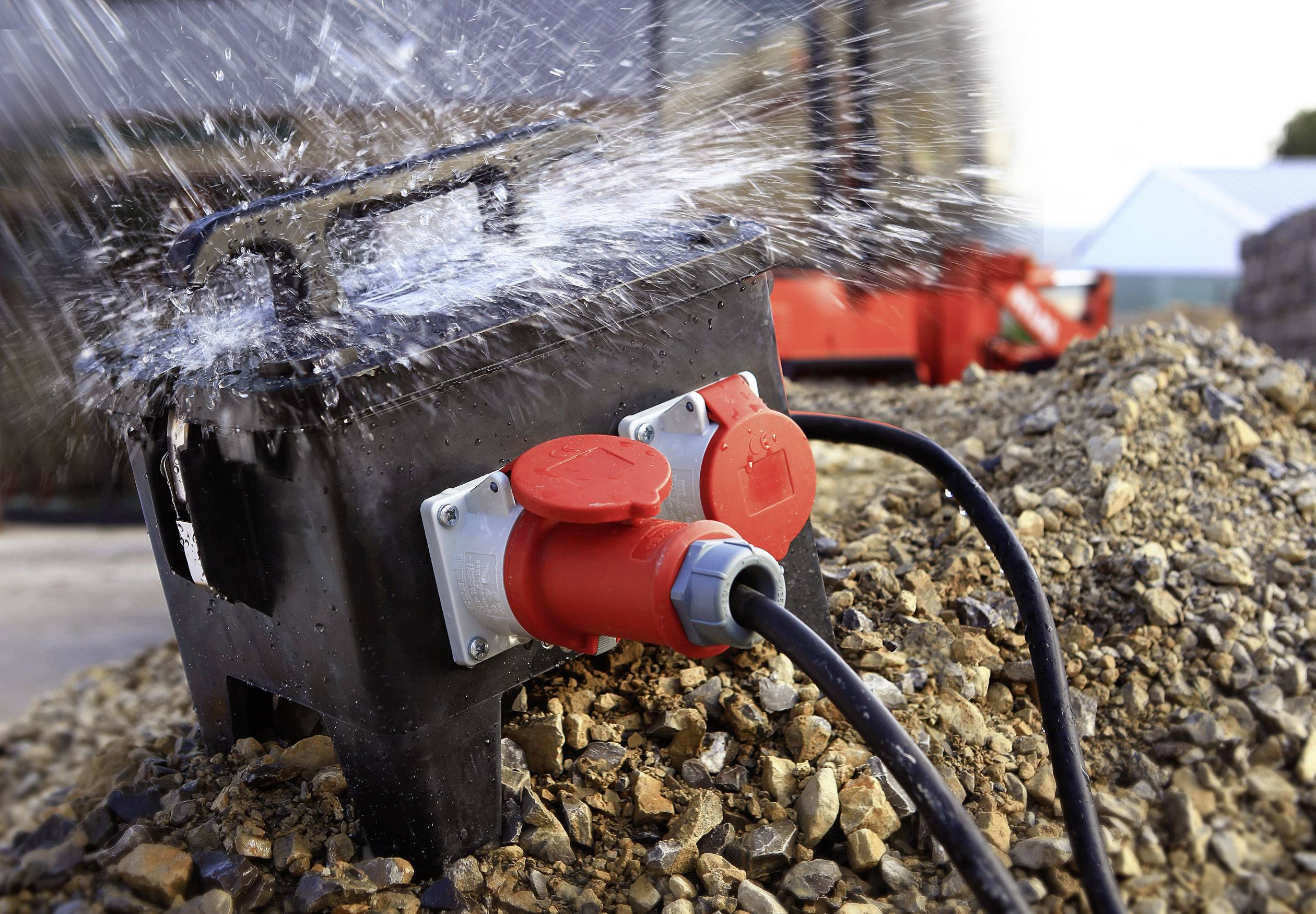 Schwarzer, spritzwassergeschützter CEE Stromverteiler mit zwei roten CEE-Steckdosen auf Schotter, ideal für die Baustelle.