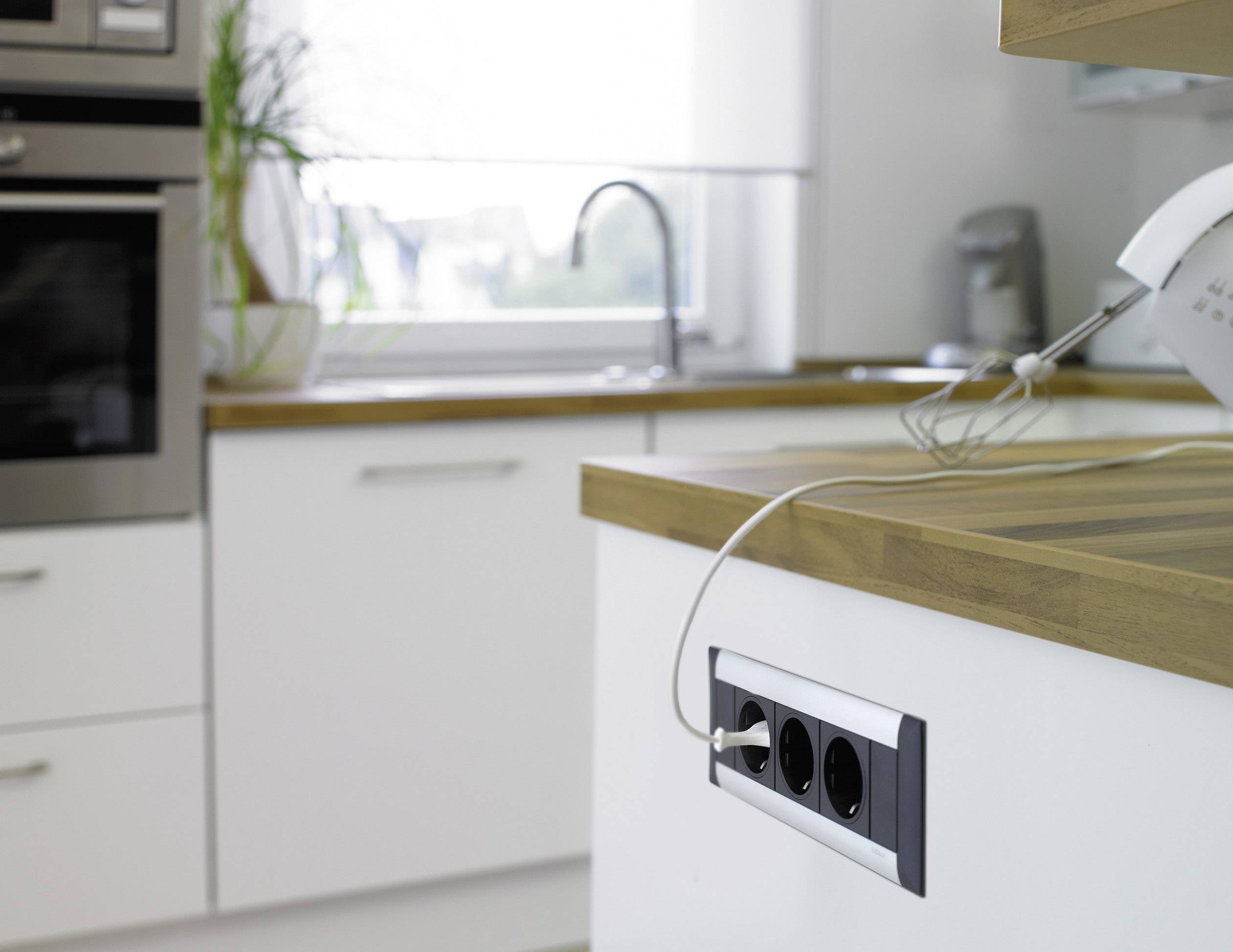Ein moderner Küchenraum mit weißem Schrank und Holzarbeitsplatte, ein Wasserkocher in einer Steckdose, Fenster im Hintergrund.