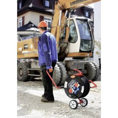 Arbeiter auf Baustelle, trägt orangenen Helm und blaue Jacke, zieht Kabeltrommel. Im Hintergrund Bagger und Gebäude.