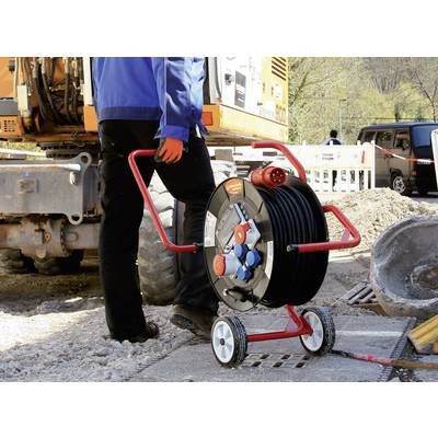 Ein Bauarbeiter schiebt eine fahrbare Kabeltrommel auf einer Baustelle. Im Hintergrund ist Baumaschinerie sichtbar.