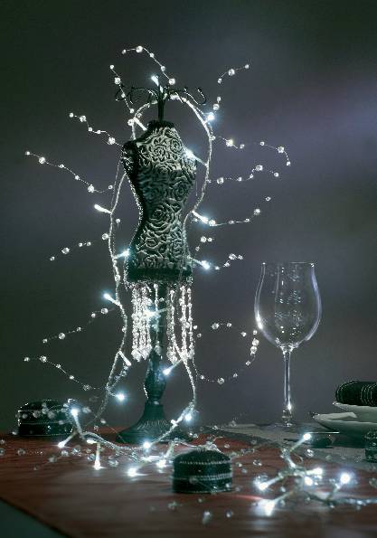 Schmuckständer in Form einer Schneiderpuppe, umhüllt von Lichterketten. Daneben steht ein leeres Weinglas auf einem roten Tuch.