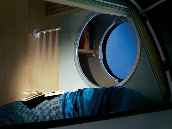 Ein gemütliches Wohnwagen-Interieur bei Nacht: Ein offenes Buch neben einer Lampe auf einem Sitz, blaues Licht kommt durch ein Bullauge.