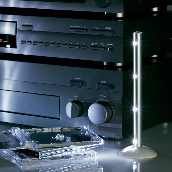 Hi-Fi-Stereoanlage mit mehreren Komponenten, zwei CD-Hüllen vorne. Rechts eine schlanke, modern gestaltete Stehlampe.