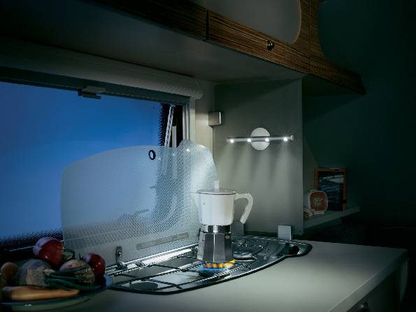 'Eine beleuchtete Wohnmobilküche mit blauem Fenster im Hintergrund. Eine Kaffeemaschine steht auf einem Gasherd, umgeben von Gemüse.'