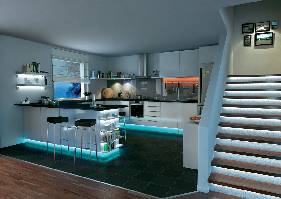 Moderne Küche mit beleuchteter Kücheninsel, Hockern und Treppe im Vordergrund. Wände sind hell, Boden in dunkler Fliese.