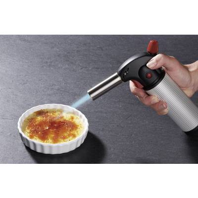 Hand hält Lötlampe über einer Crème Brûlée in einer weißen Schale, während die Oberfläche karamellisiert wird.